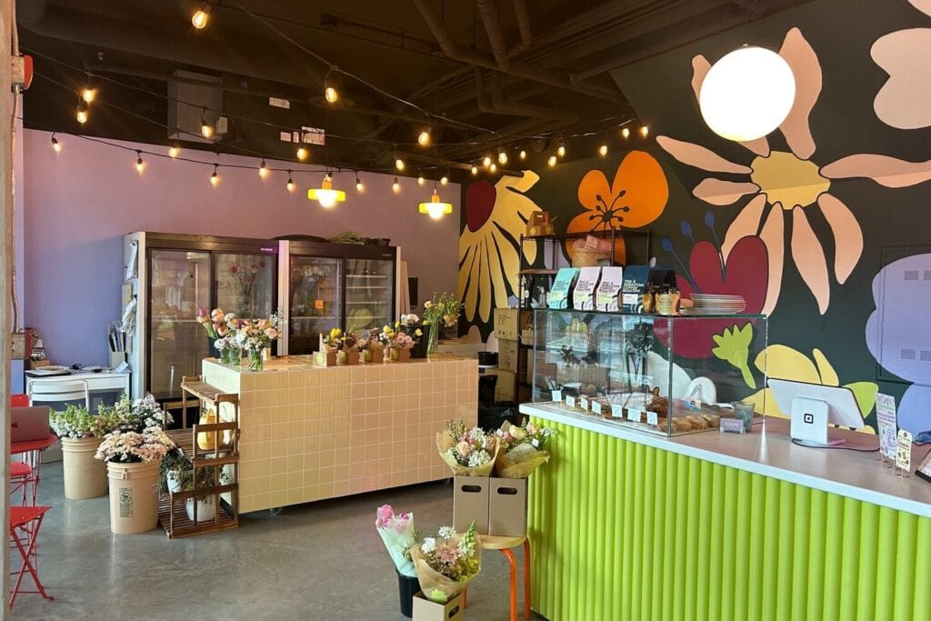 Café fleuri avec comptoir vert, fresque murale colorée et présentoirs réfrigérés.