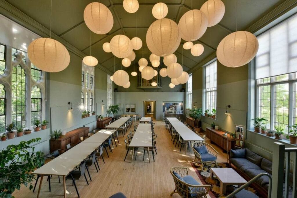 : Grande salle de réception lumineuse avec longues tables, plantes et lanternes suspendues.