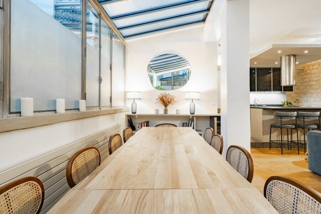 Grande table de travail en bois sous une verrière, à côté d'une cuisine ouverte.
