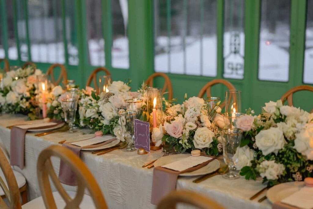 Longue table de réception fleurie dans une orangerie vitrée éclairée par des lustres.