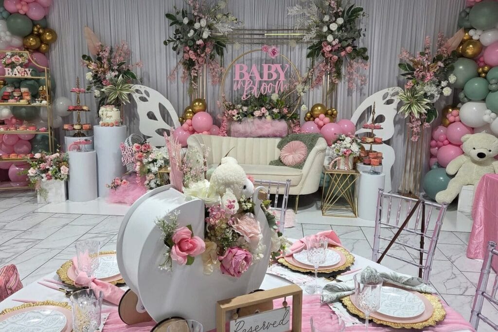 Décor rose et or pour une baby shower avec ballons, fleurs, fauteuil et tables dressées.