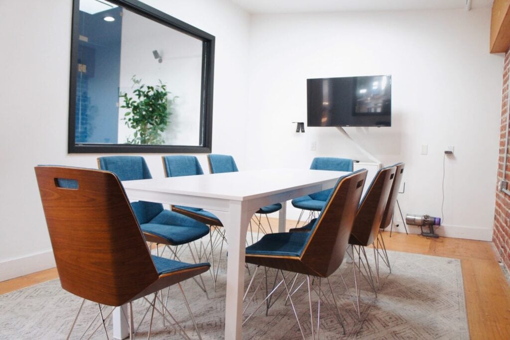 Salle de réunion fermée avec grande table, chaises bleues, écran mural et mur en briques.