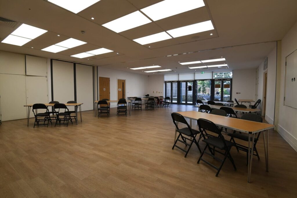 Salle polyvalente avec tables, chaises pliantes, parquet clair et accès direct sur l'extérieur.