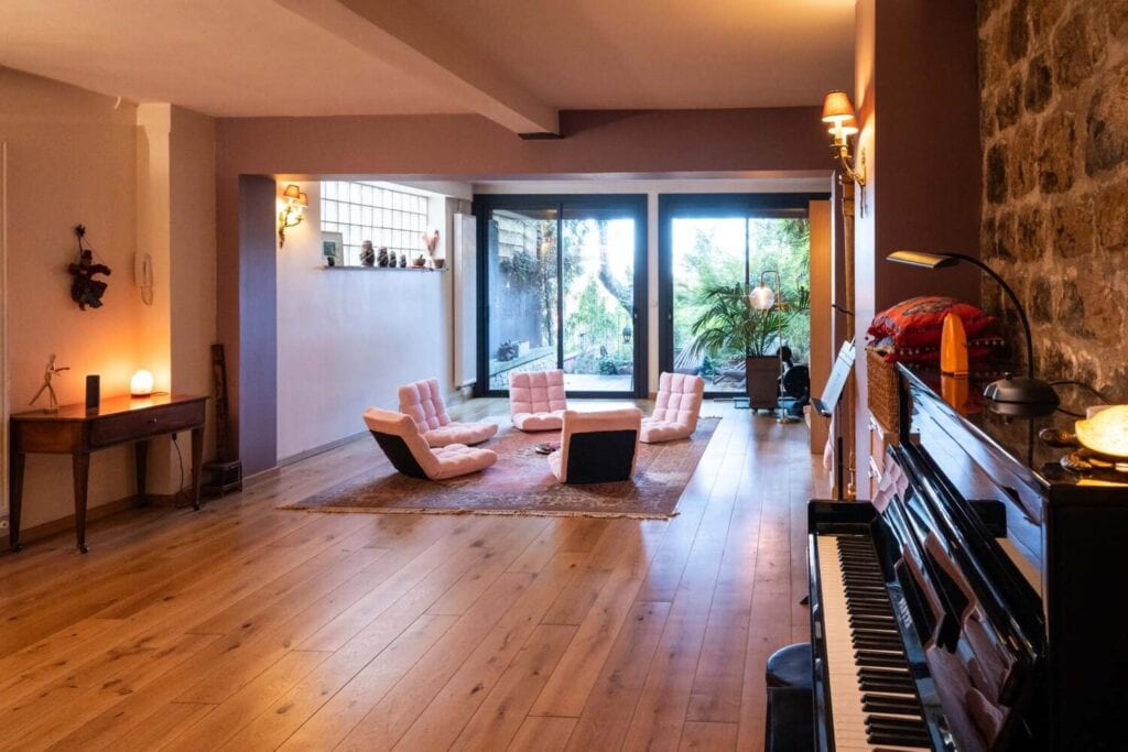 Salle polyvalente lumineuse avec parquet, piano et baies vitrées ouvertes sur un jardin.