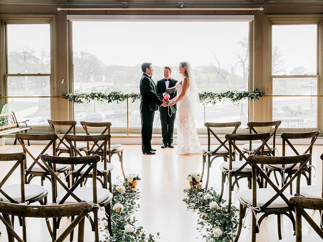 Couple being officiated in front of large windows and empty chairs at NYC wedding venue