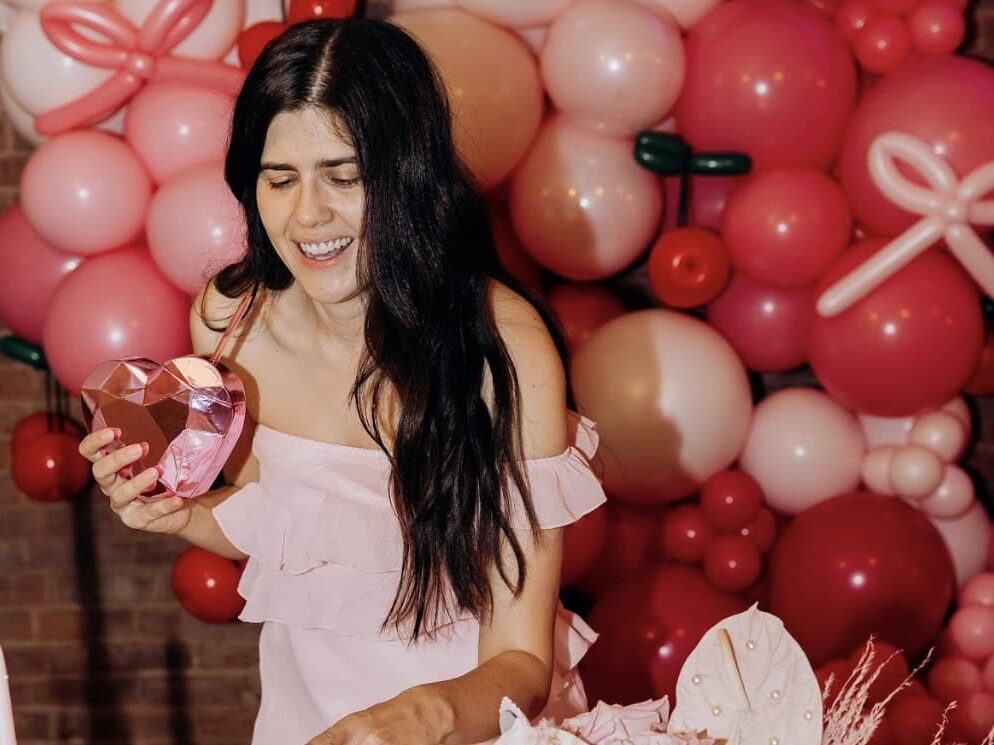 Girl wearing pink Galentines outfit in front of pink and red balloons cutting pink heart cake