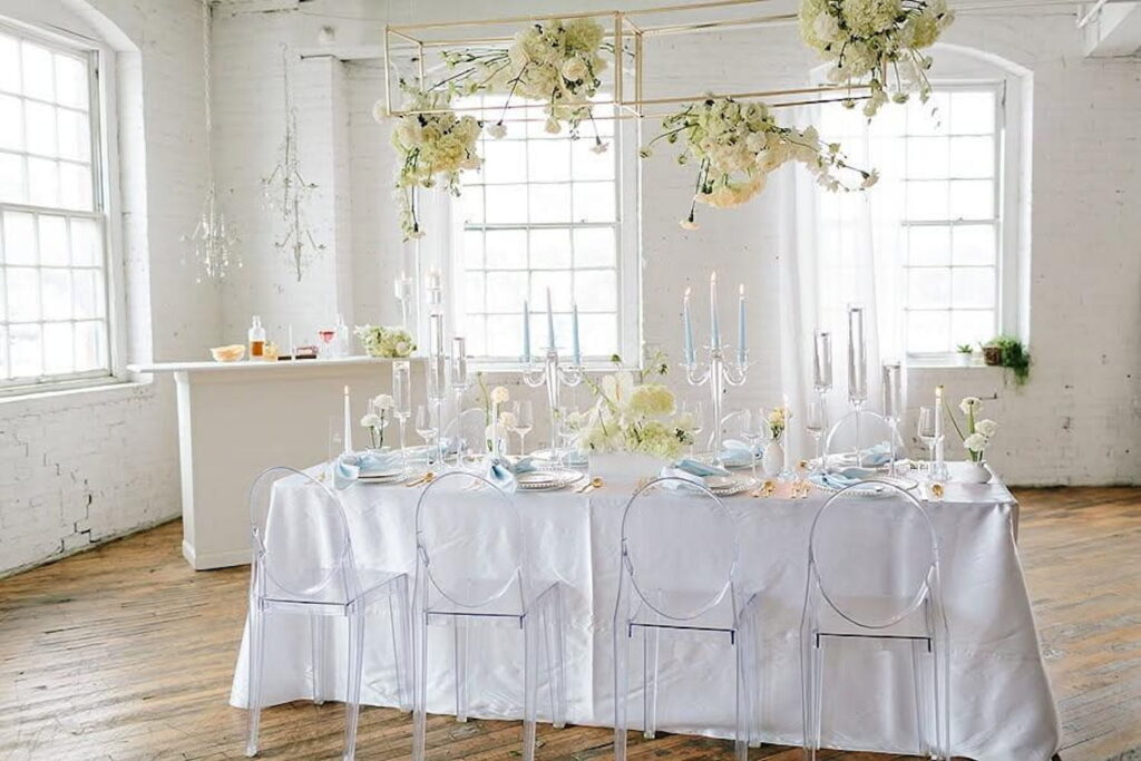 All-white brick event space with small bar in corner, large paned windows, and a small banquet table draped in white with blue accents