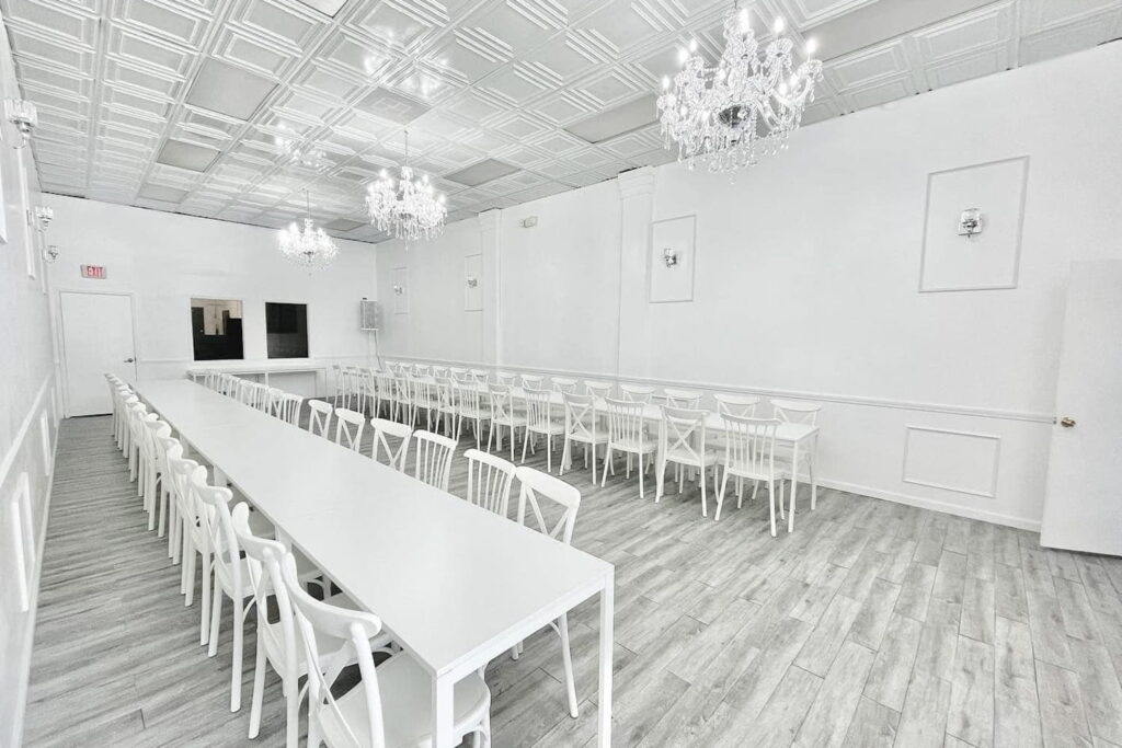 All white event space with long white tables and chairs and chandeliers hanging from ceiling