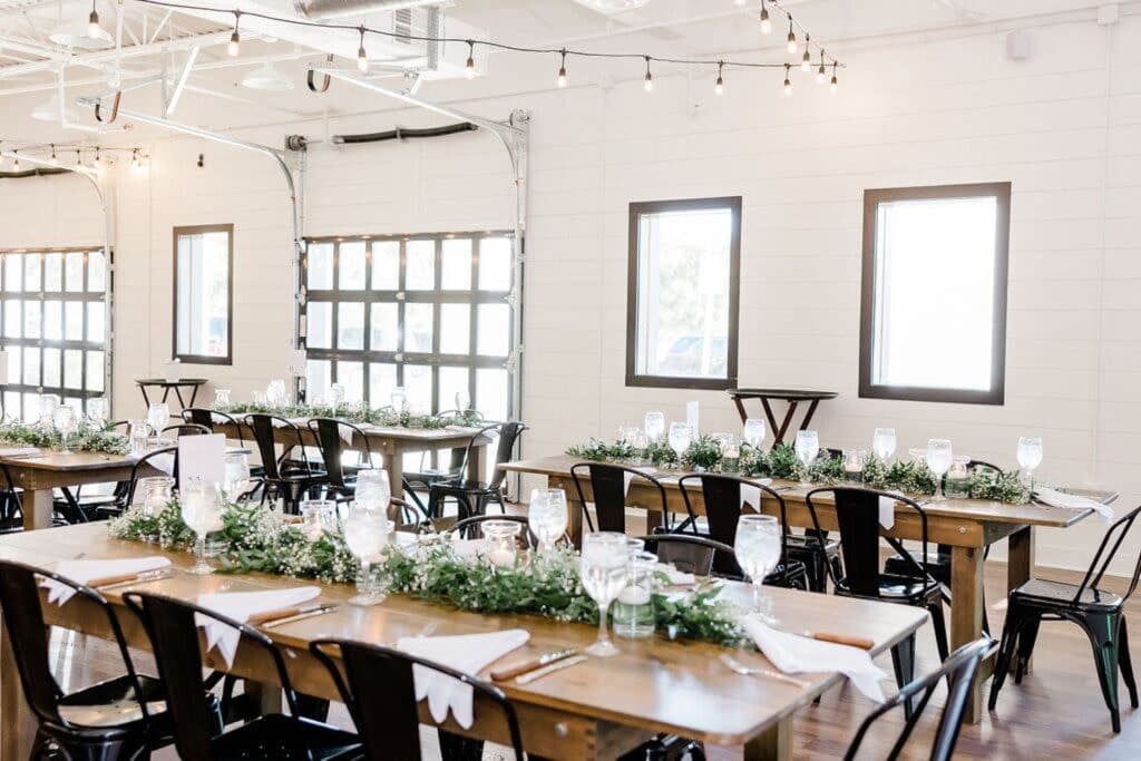 A modern event venue with white walls and black garage doors and windows is set up with wooden tables decorated in garlands of greenery, black metal chairs and low candles