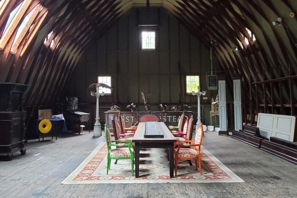 A long table on a red and cream rug surrounded by brightly colored chairs in a very tall barn with soaring ceilings