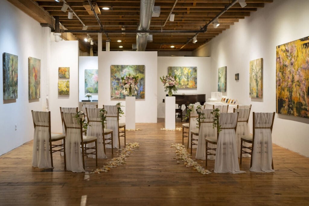 A small wedding ceremony is set up in an art gallery with white walls and floral paintings on each wall
