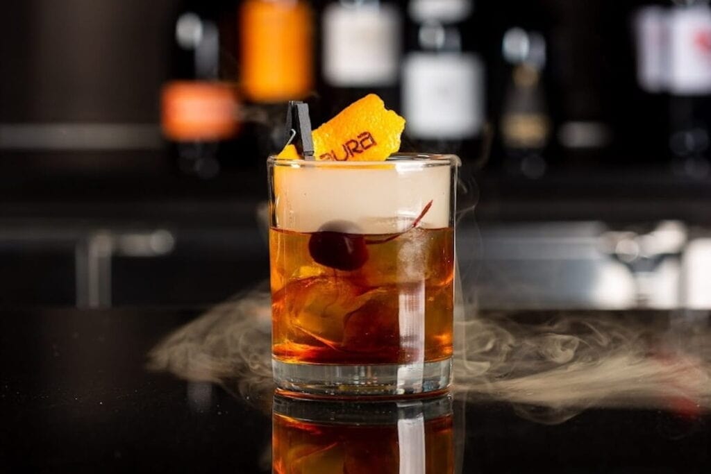 A richly colored cocktail sits on a bar surrounded by a swirl of smoke