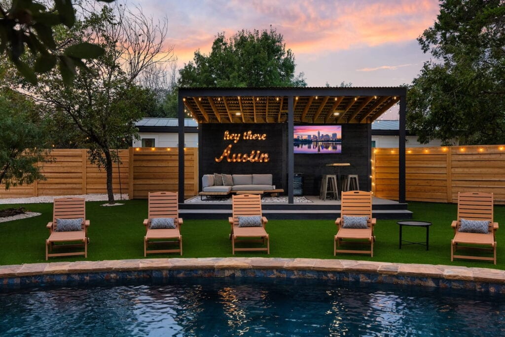 Lounge chairs sit by a pool's edge in a backyard with a covered seating area housing a couch, big screen tv, and a light up sign that says "Hey There Austin"