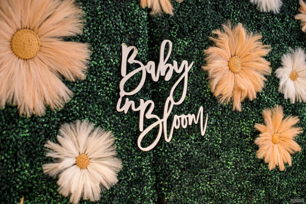 A "Baby in Bloom" sign hangs on a wall made of greenery and decorative flowers