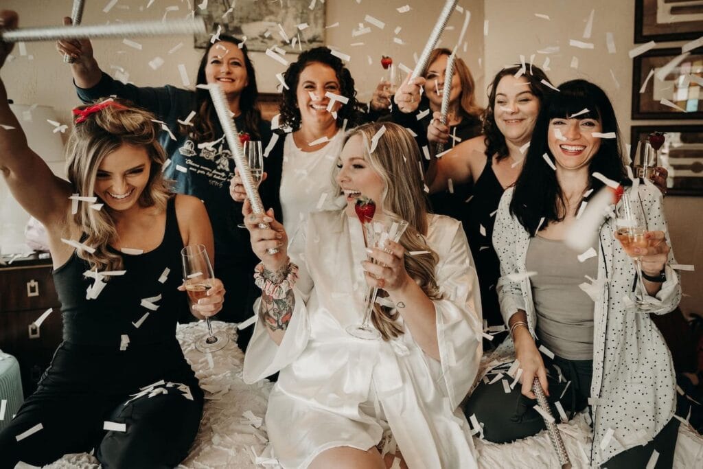 A group of women dressed in black and white surround a bride to be in a white robe as confetti floats down from above