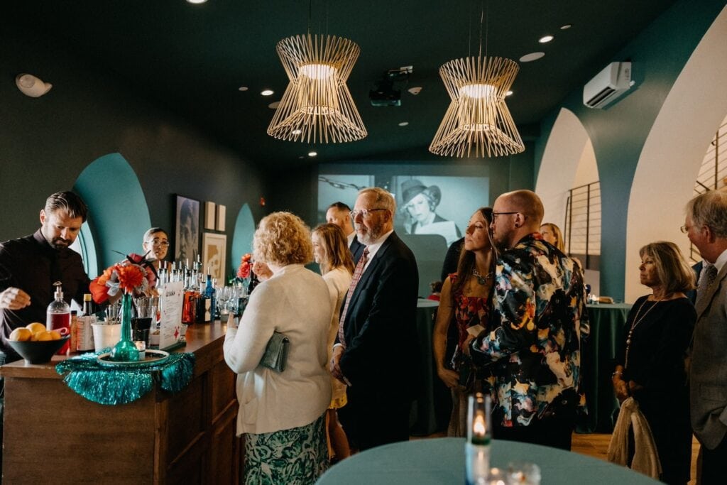 Party goers line up at a bar in a moody, dark green venue space punctuated by a black and white movie projected on a wall and gold decor throughout