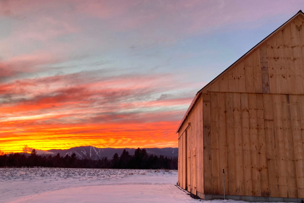 A wood barn sits in the foreground with a fiery sunset in the background above mountains