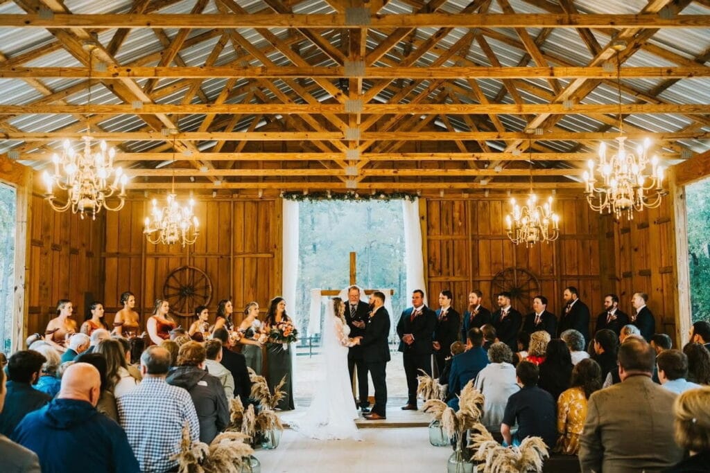 A couple exchanges vows at an altar in a rustic barn with dramatic chandeliers hanging from wooden beams in front of many friends and family