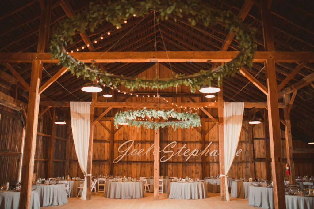 A large barn is set up for a wedding reception with greenery and string lights hanging from beams and a giant sign on the wall of the bride and groom's names.