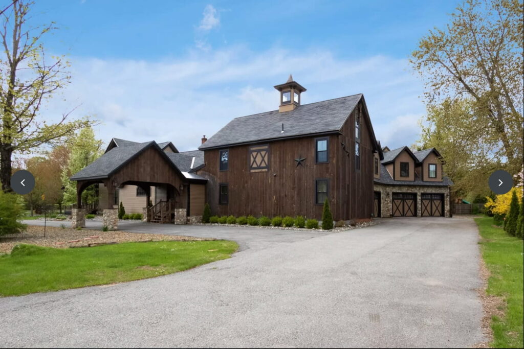 A large, dark wooden barn attached to a three car garage set on a circular drive surrounded by trees