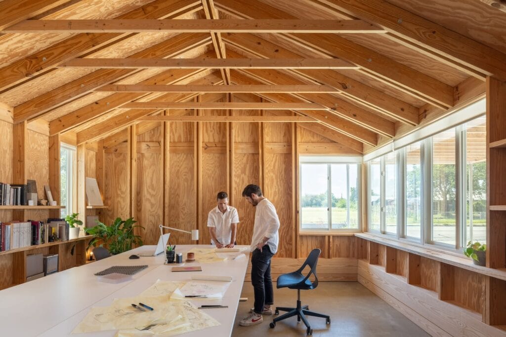 Two men stand at a work table reviewing building plans in a workspace with exposed wood beams, high ceilings and lots of windows
