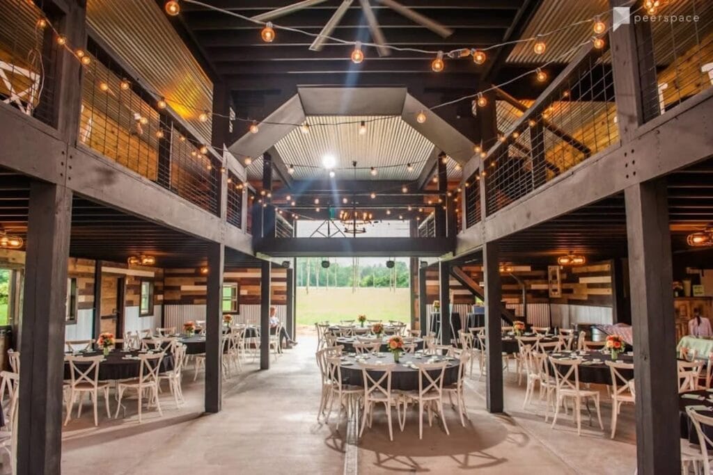 A large wood barn with corrugated metal ceilings strung with bistro lights is set up with round tables and white chairs for a wedding reception