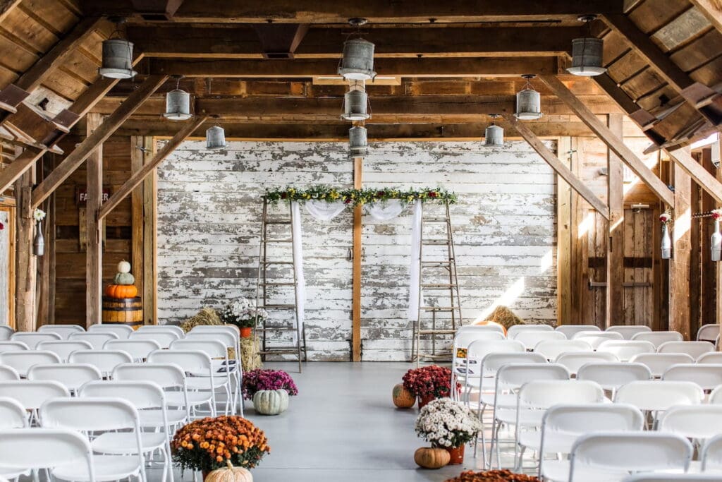A rustic barn is set up for a wedding ceremony, decorated with pumpkins and mums