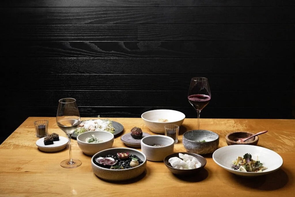 A selection of plated dishes and glasses of wine on a light wood table in front of a dark shiplap wall