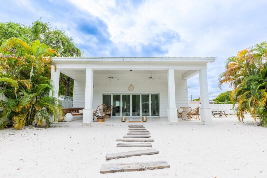 An all-white beachfront home sits in the sand surrounded by palm trees