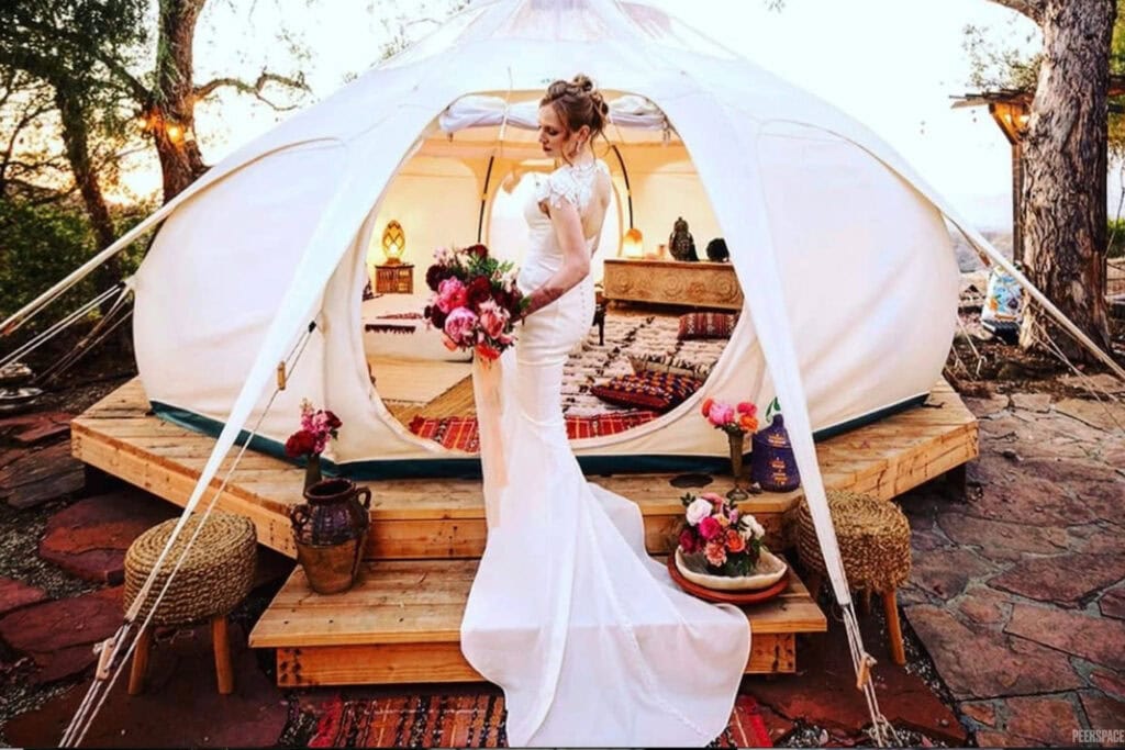 A bride stands on the stairs of a beautifully-decorated yurt outside while the sun sets in the distance