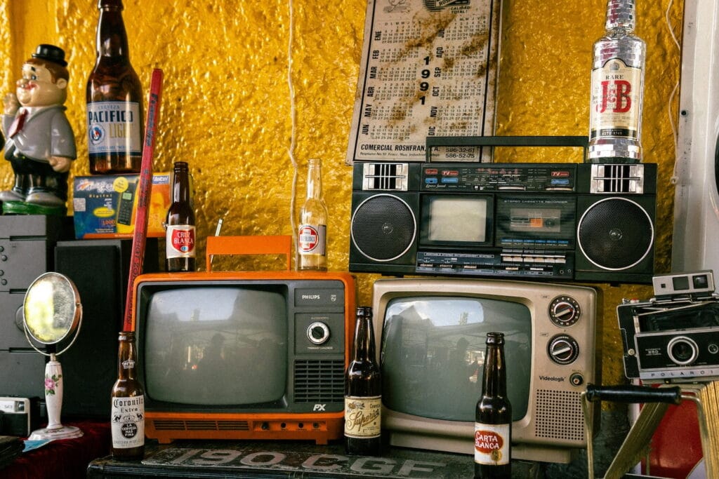 Old TVs, cameras and radios surrounded by old beer bottles