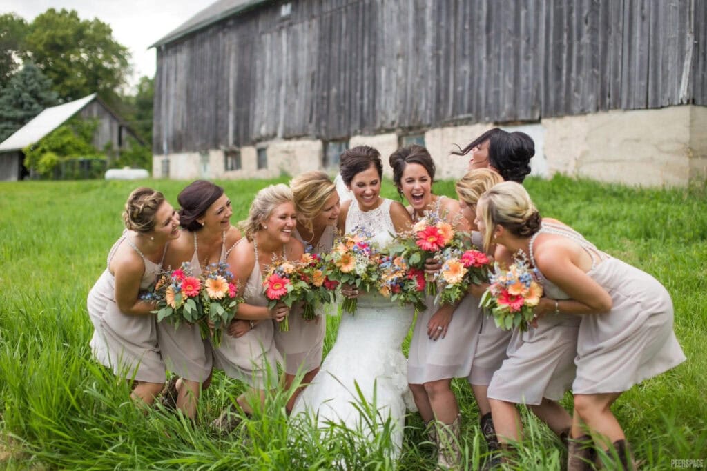 A bridal party poses while laughing with the bride in front of a large barn