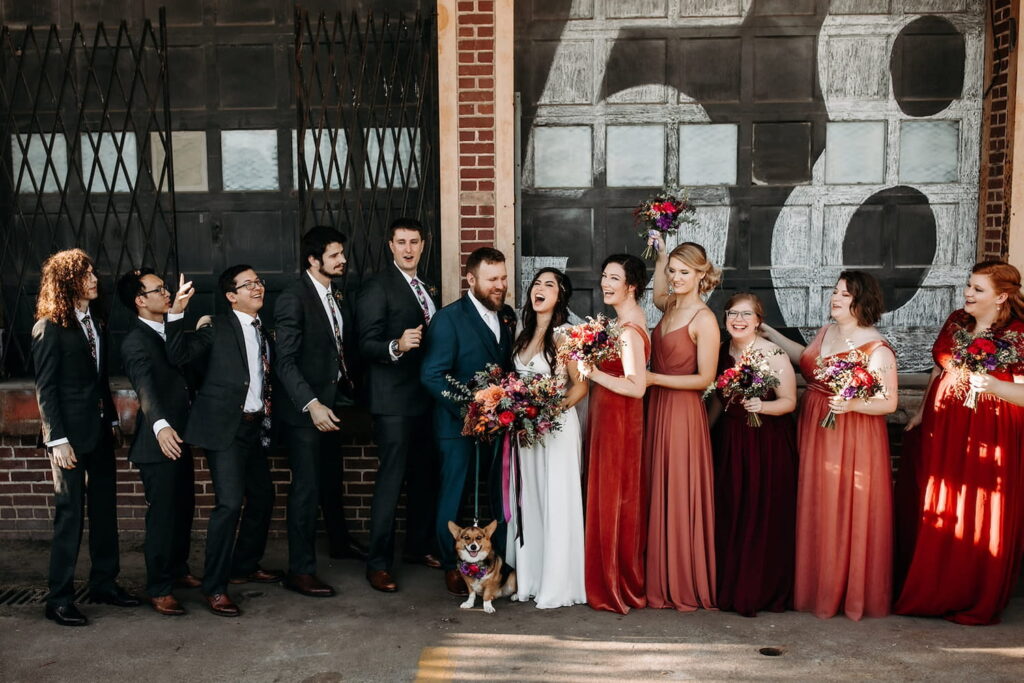 A bride and groom stand outside of a brick building with their dog and wedding party who are all cheering