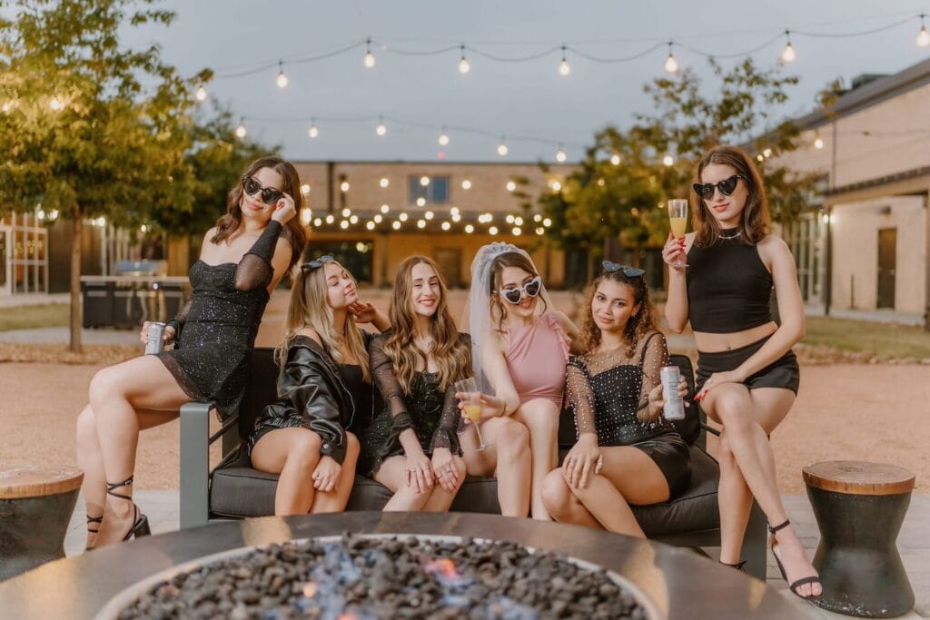 Bachelorette party sitting on outdoor couch under string lights wearing heart-shaped sunglasses
