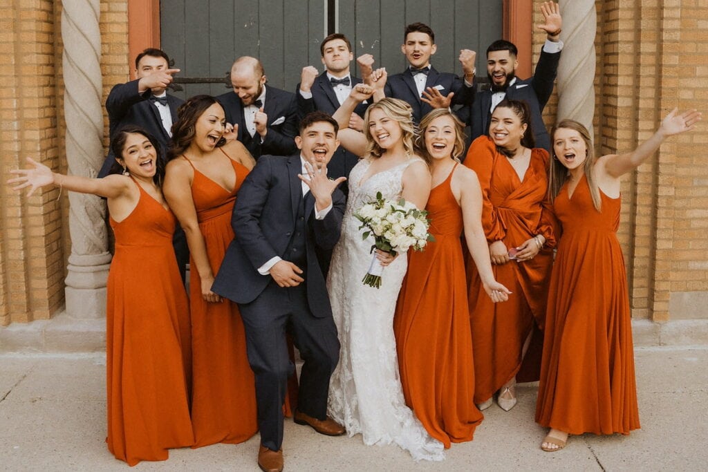 Bridesmaids in orange dresses and groomsmen in black tuxes surround a bride and groom outside of a brick building