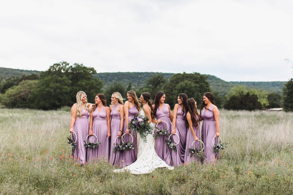 Bridesmaids in lavender dresses stand with a bride in an open field