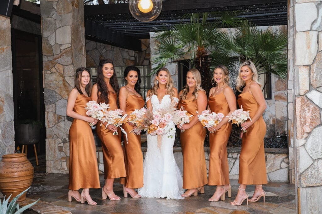 Bridesmaids in burnt orange dresses stand next to a bride on a stone patio