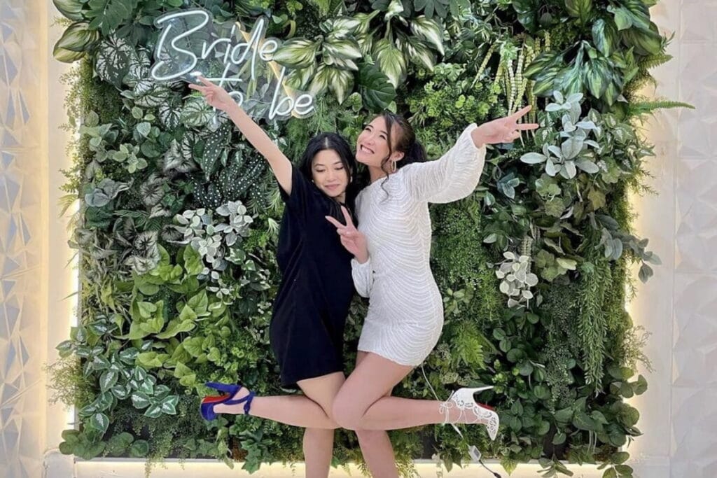Two women pose in front of a Bride to Be sign lit up against a wall of greenery
