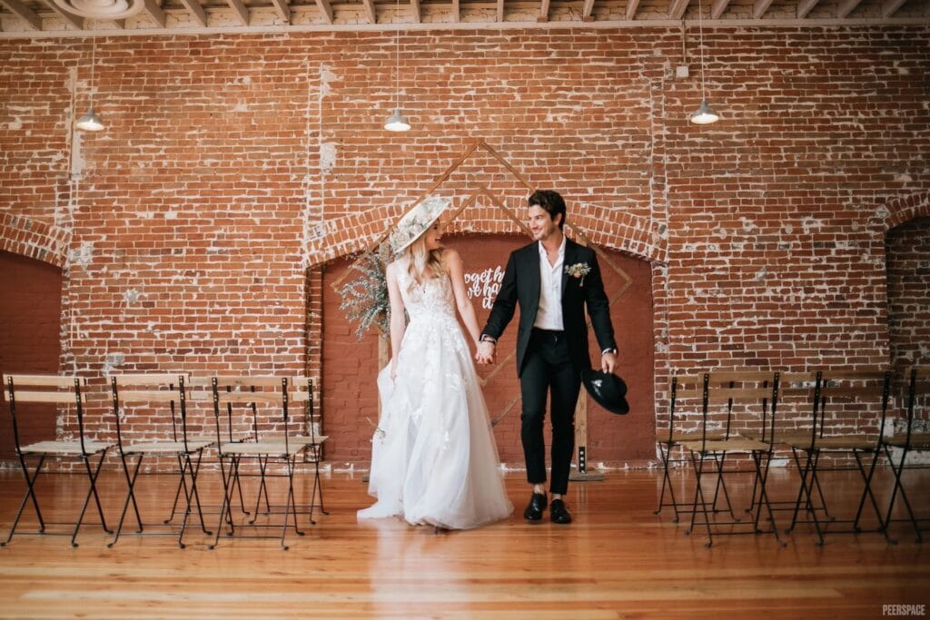 A stylish bride and groom walk hand-in-hand down aisle in industrial space with wooden geometric altar