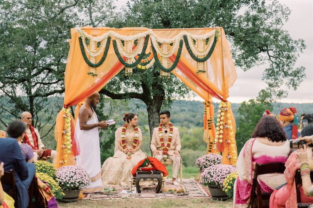 A bride and groom sit outdoors under a traditional orange and yellow mandap