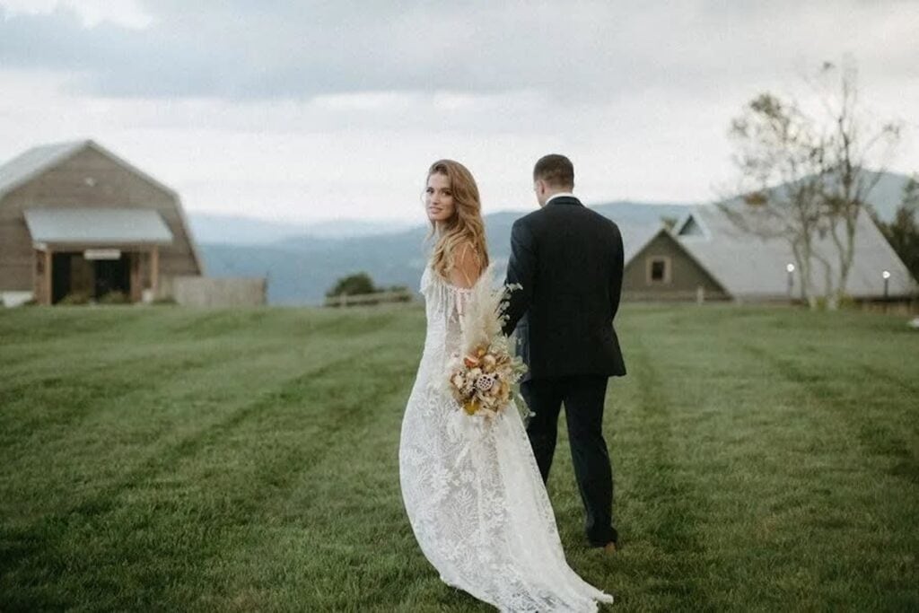 A bride and groom walk away from the camera in the grass towards a barn while the bride looks back smiling, holding a bohemian-inspired bouquet
