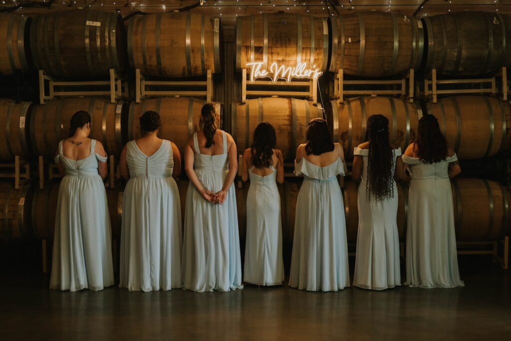 A bride and her bridal party pose in front of a wall of wine barrels while facing away from the camera