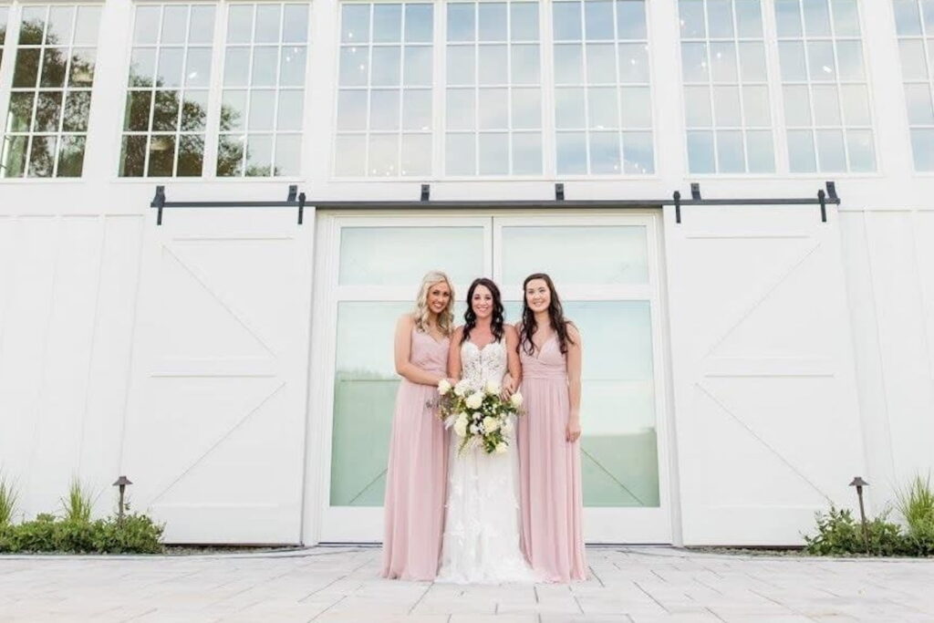 A bride with two bridesmaids dressed in pink stand outside giant white barn doors