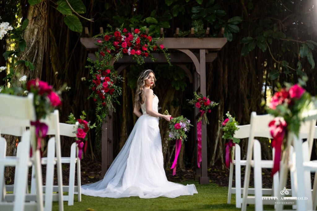 A bride looks back from the front of an aisle by a wooden altar draped in bright red flowers with wooden ceremony chairs on either side