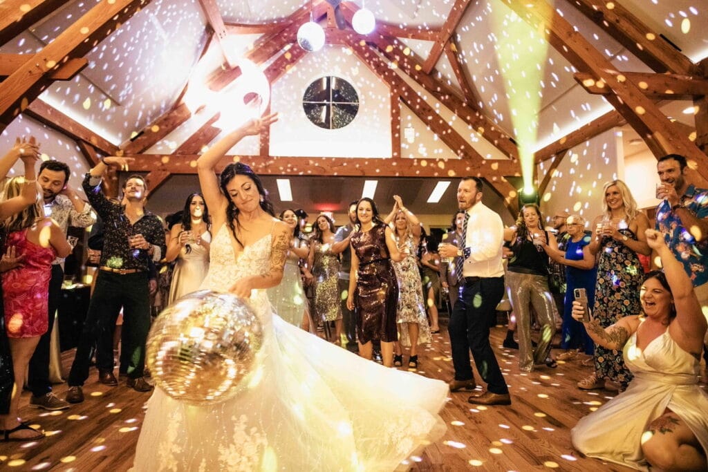 A jubilant crowd surrounds a bride on a dance floor as she raises a peace sign and swings a disco ball