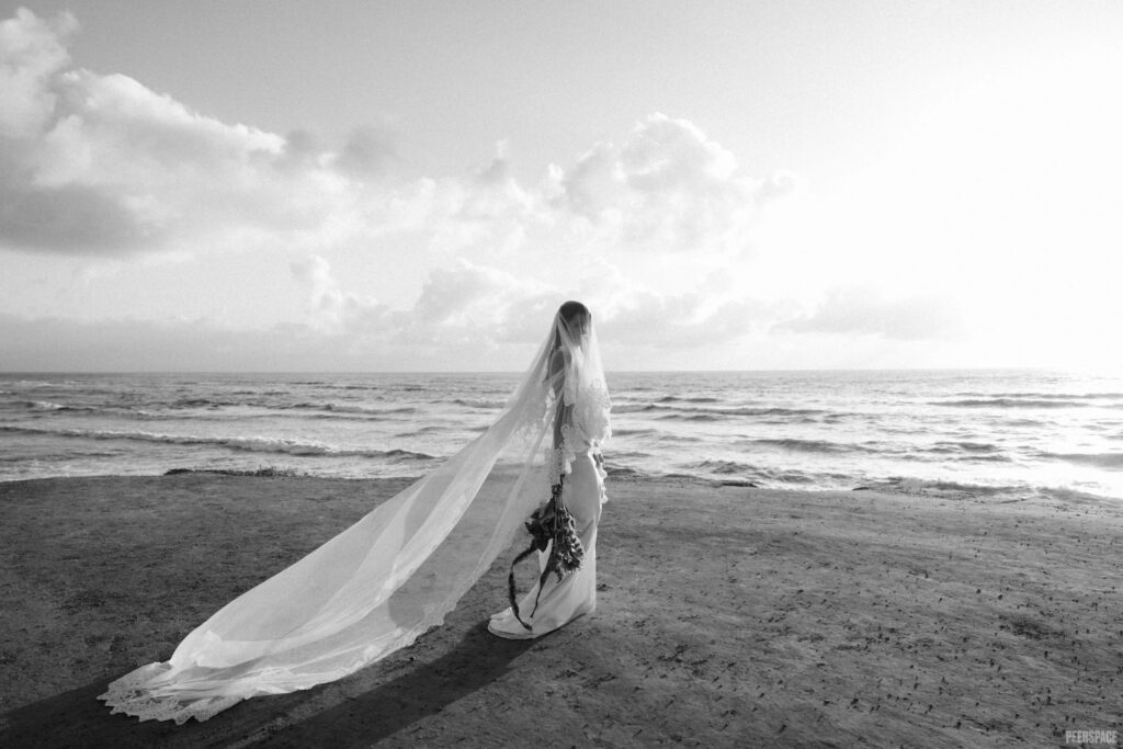 A bride draped in a dramatic veil stands on the beach holding bouquet