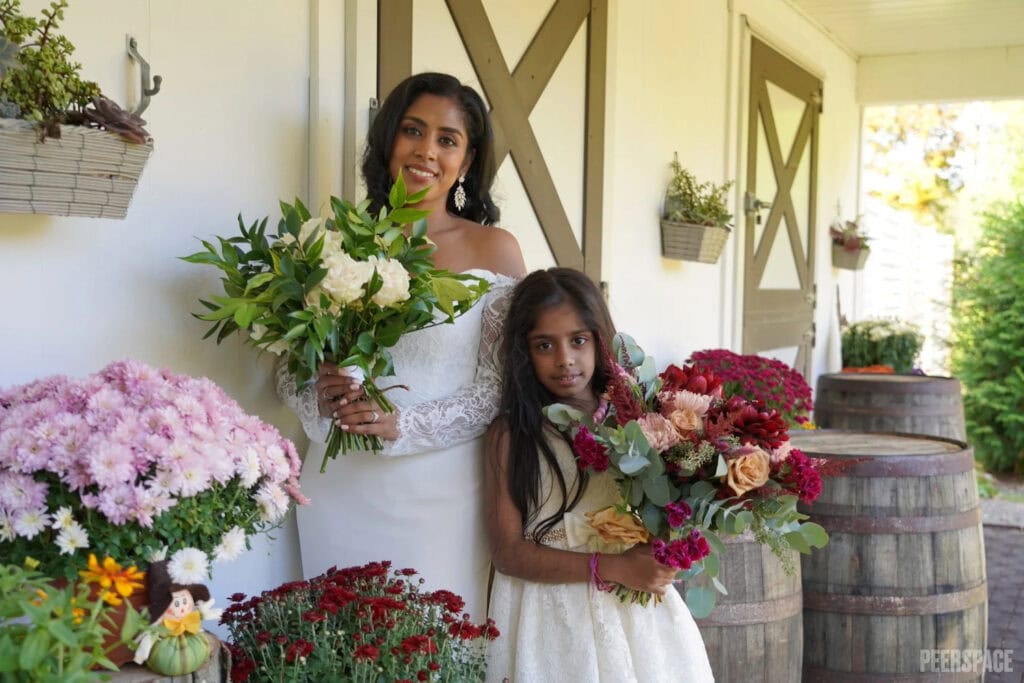 A bride and a flower girl pose with bouquets outside a rustic barn