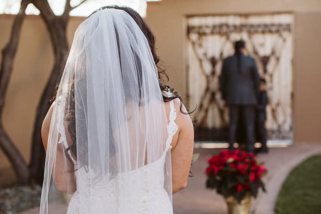 A bride is in the foreground, photographed from the back, looking at her groom in the background standing by a large wooden door
