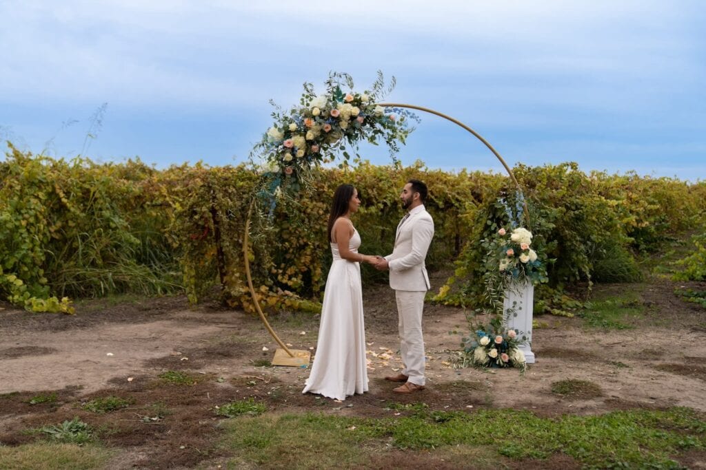 A bride and groom stand hand-in-hand in front of a round altar decorated in pink, white and yellow flowers in front of a farm