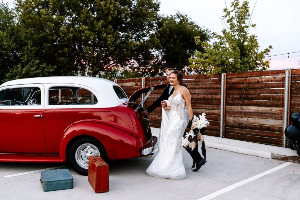 A bride and groom stand near a red and white antique car